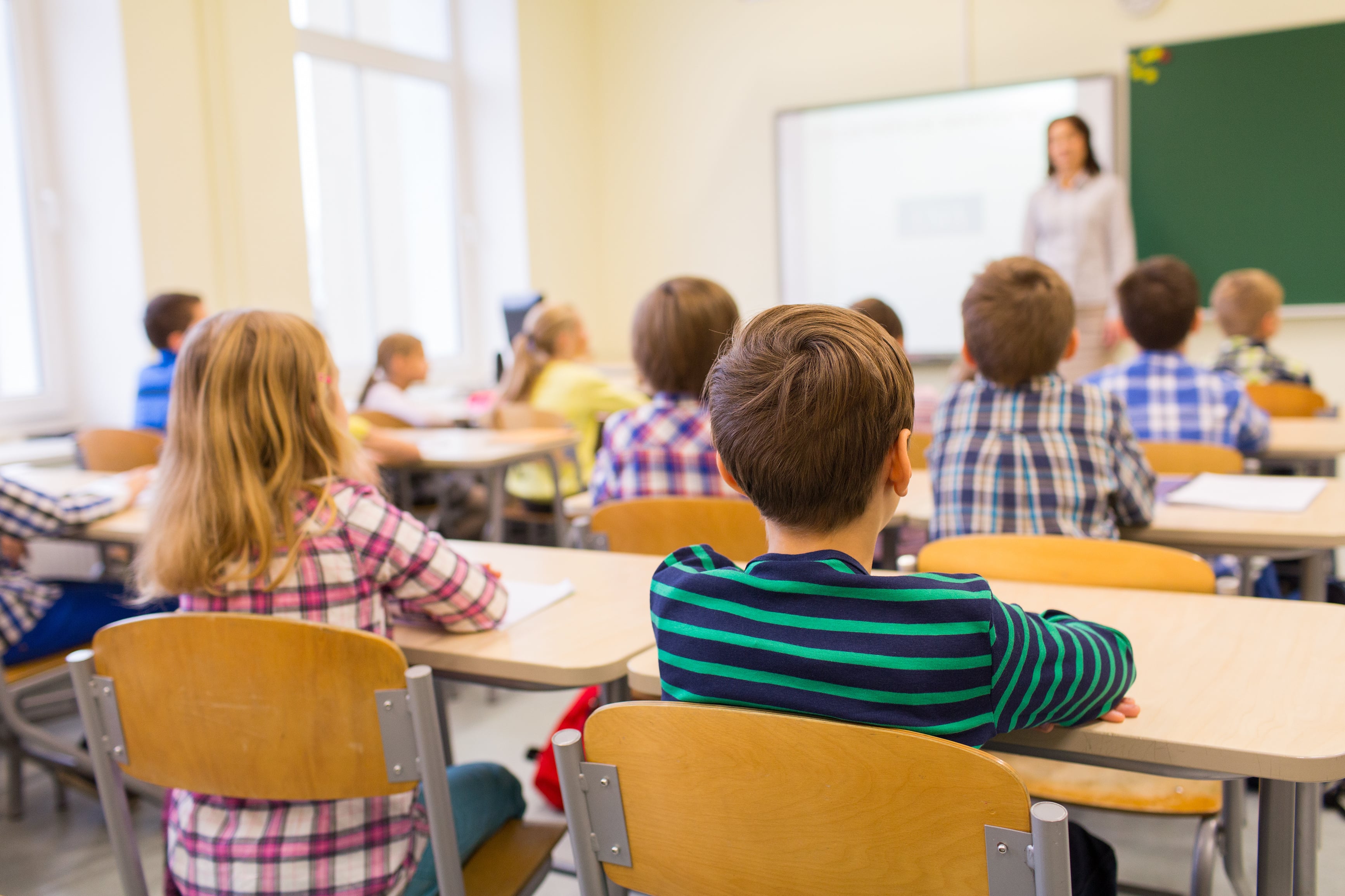 Elementary school students seated at desks in a classroom, attentively facing the teacher at the front of the room