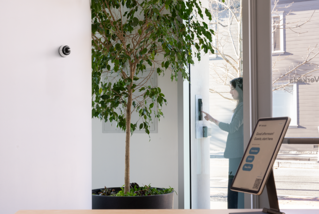 Visitor using a secure intercom access control system at office entrance, with indoor surveillance camera and digital visitor check-in kiosk visible in modern lobby.